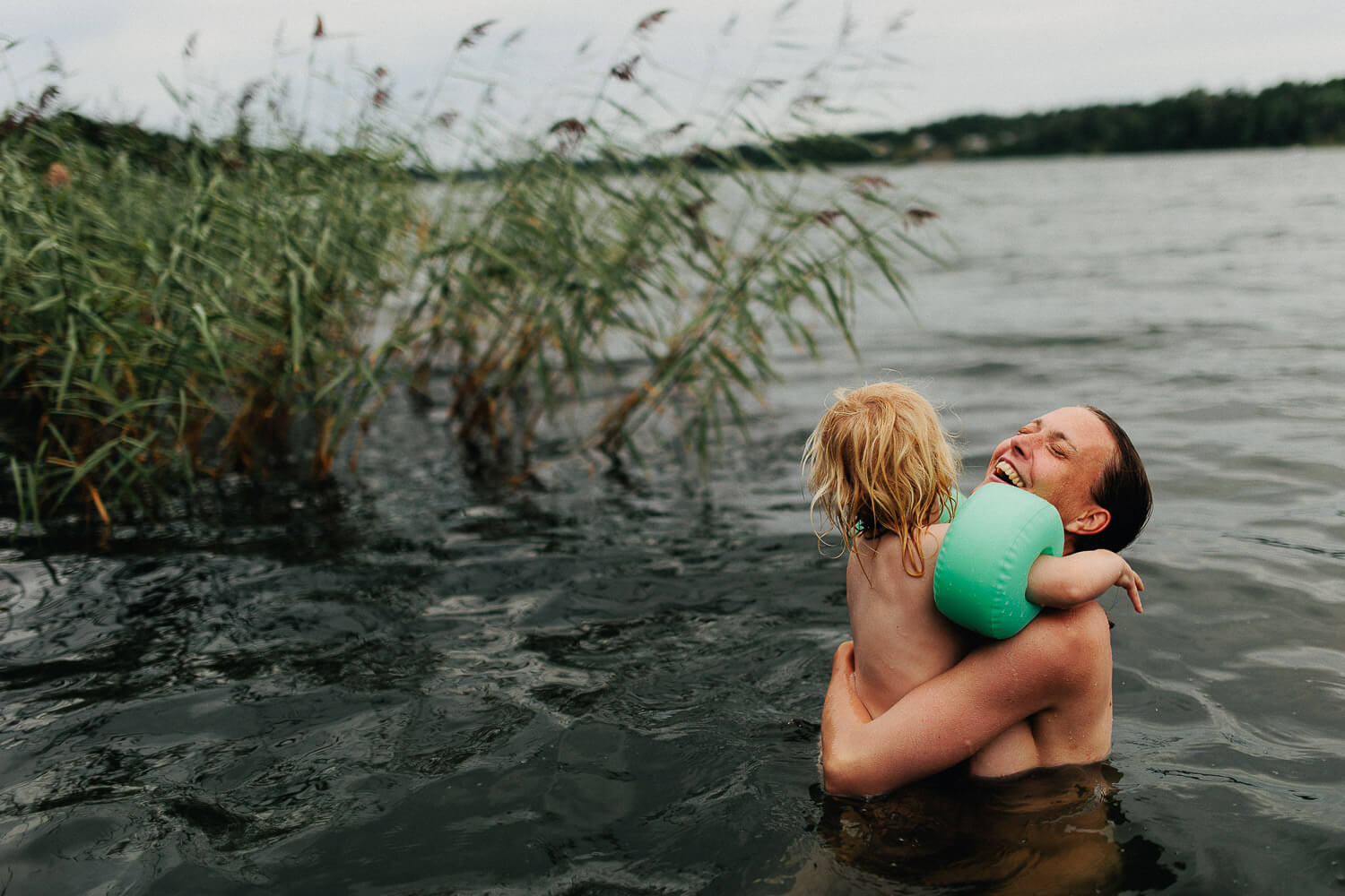 Mamma skrattar med sin dotter i famnen under en familjefotografering vid havet.